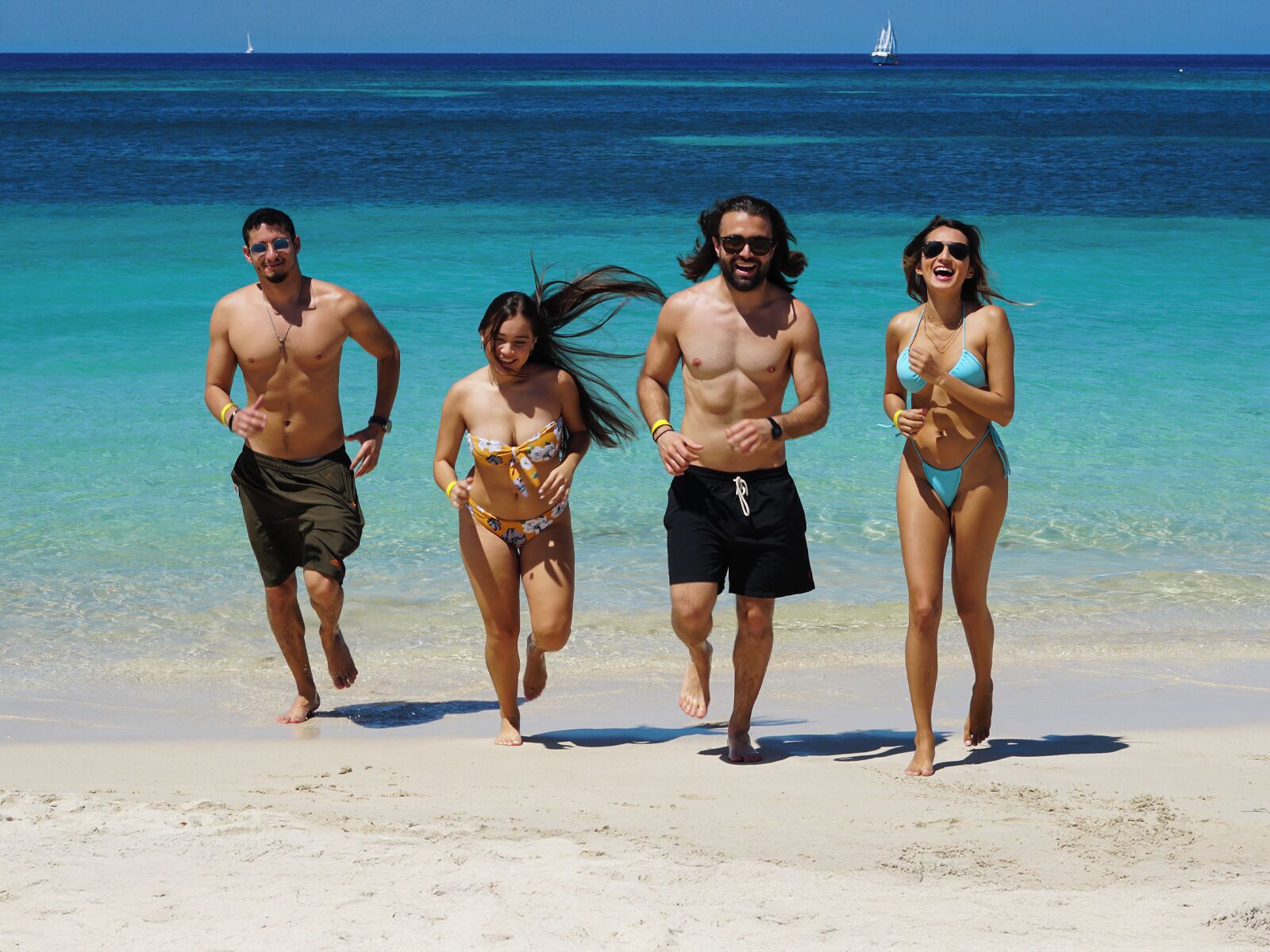 Escena de playa con agua turquesa cristalina y un velero a lo lejos.