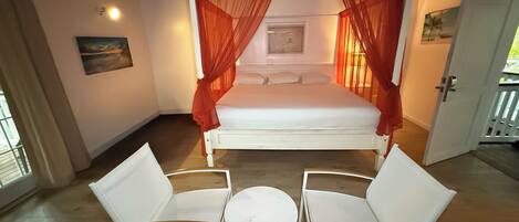 A bedroom with a canopy bed, two white chairs, a small round table, and a painting on the wall.