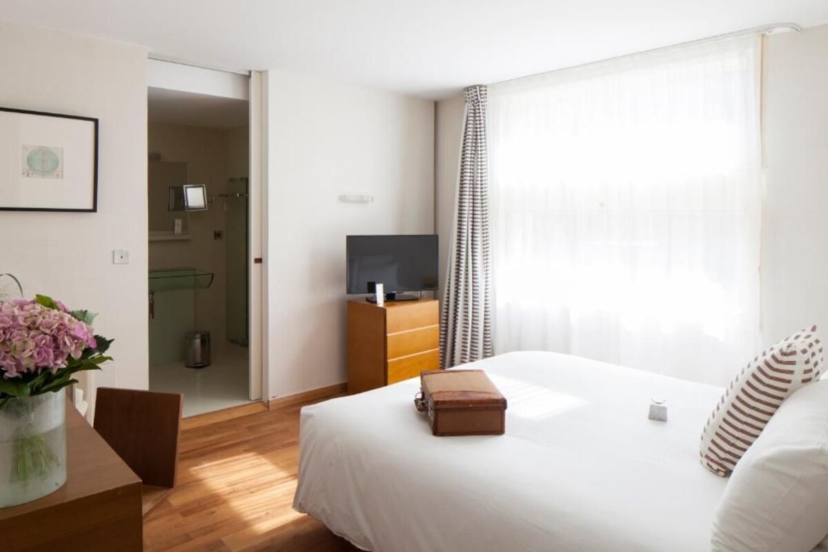 Habitación de hotel con una cama, un televisor sobre un tocador de madera, un jarrón con flores y una puerta que da al baño.