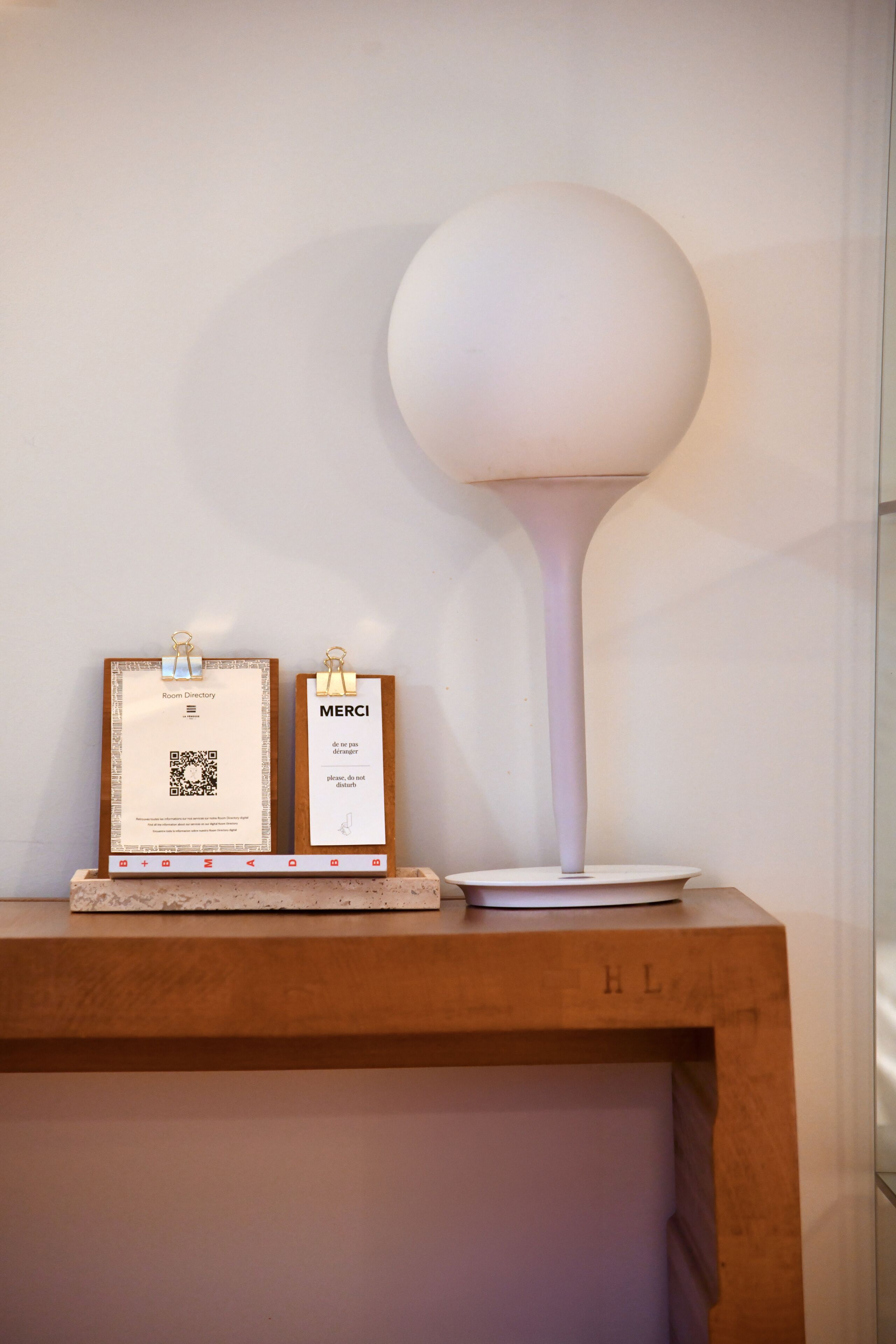 A room with a wooden shelf holding a framed document labeled 'Room Directory' and a sign with 'MERCI' and a QR code. A white lamp with a spherical top is on the shelf.