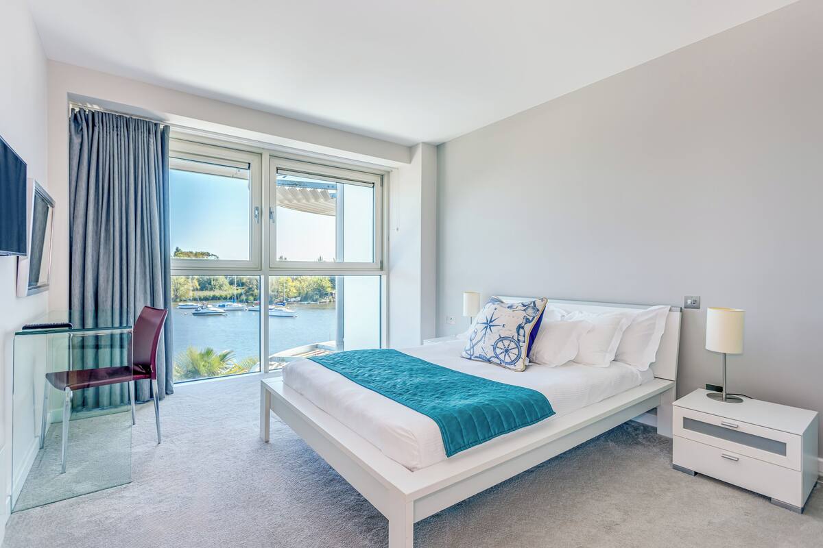A modern hotel room with a large window, a bed with white linens and a teal throw, a bedside table with a lamp, and a chair by the window.