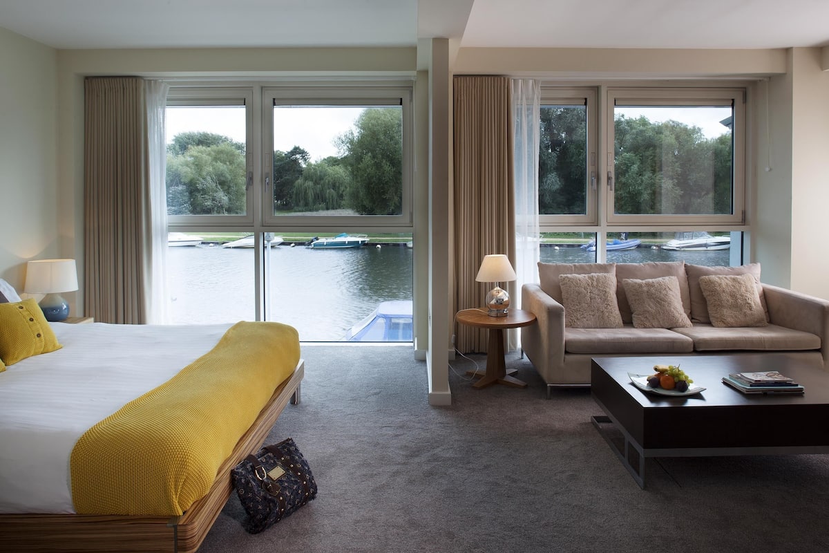 A hotel room with a bed, a sofa, a coffee table, and a view of a lake with boats.