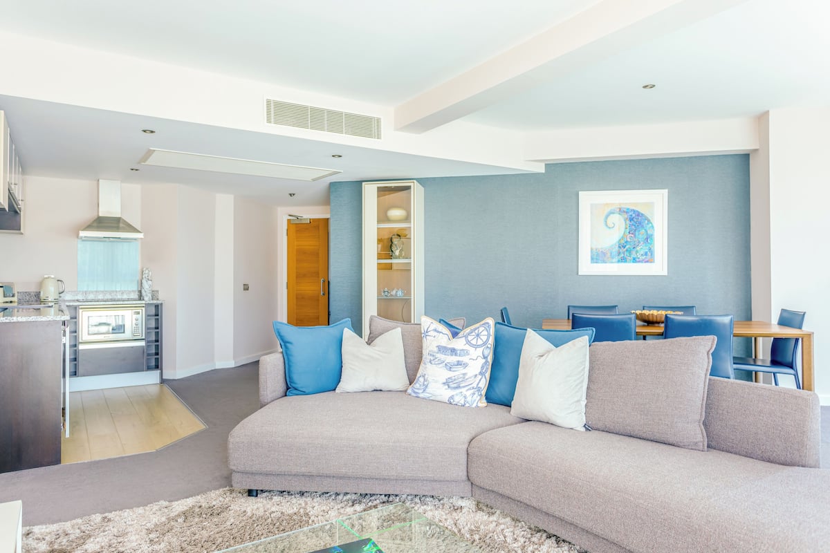A modern living room with a grey sectional sofa, a dining area with blue chairs, and a kitchen in the background.