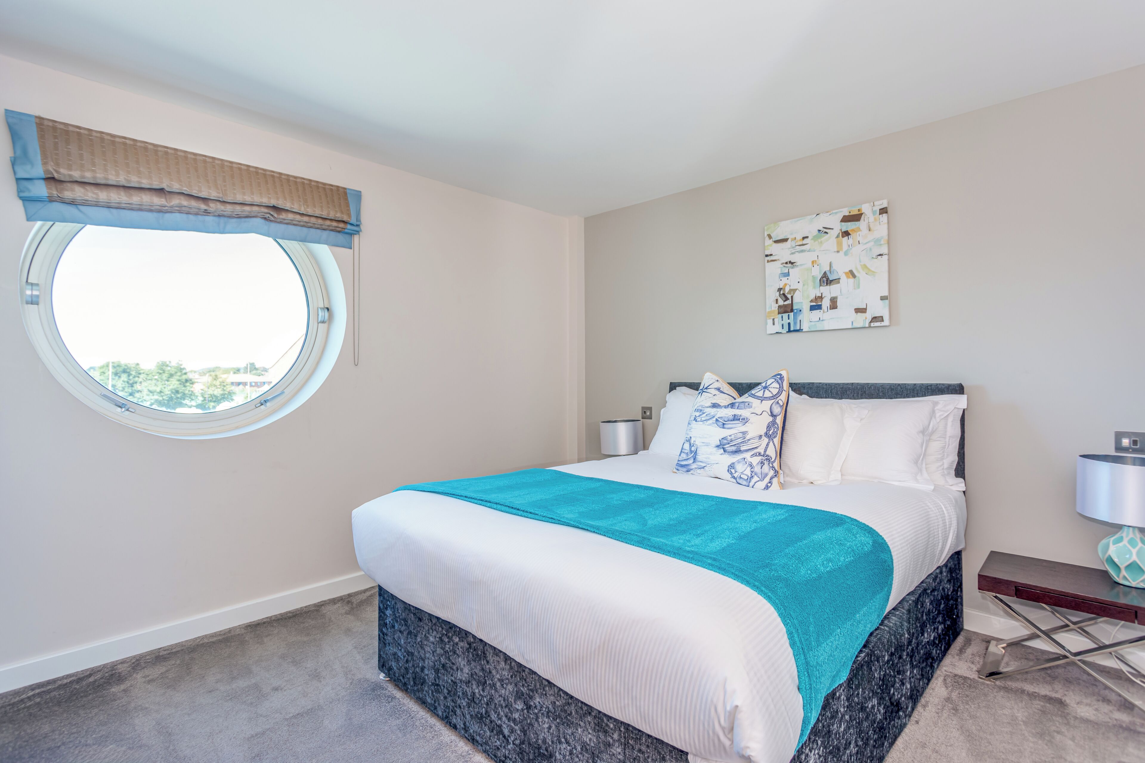A bedroom with a round window, a bed with a blue and white bedspread, a nightstand, and a wall-mounted lamp.