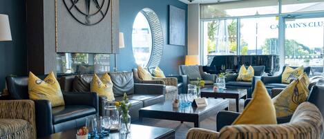 A modern lounge area with leather sofas, patterned armchairs, and a large wall-mounted compass. Tables are set with glasses and vases.