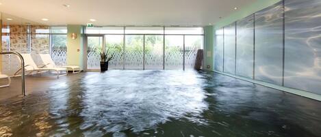 Una amplia piscina interior con agua cristalina, rodeada de sillones de descanso y un muro de cristal.