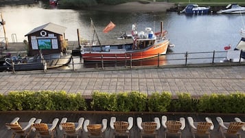 A riverside dock with boats, a small building with a lifebuoy sign, and lounge chairs along the walkway.