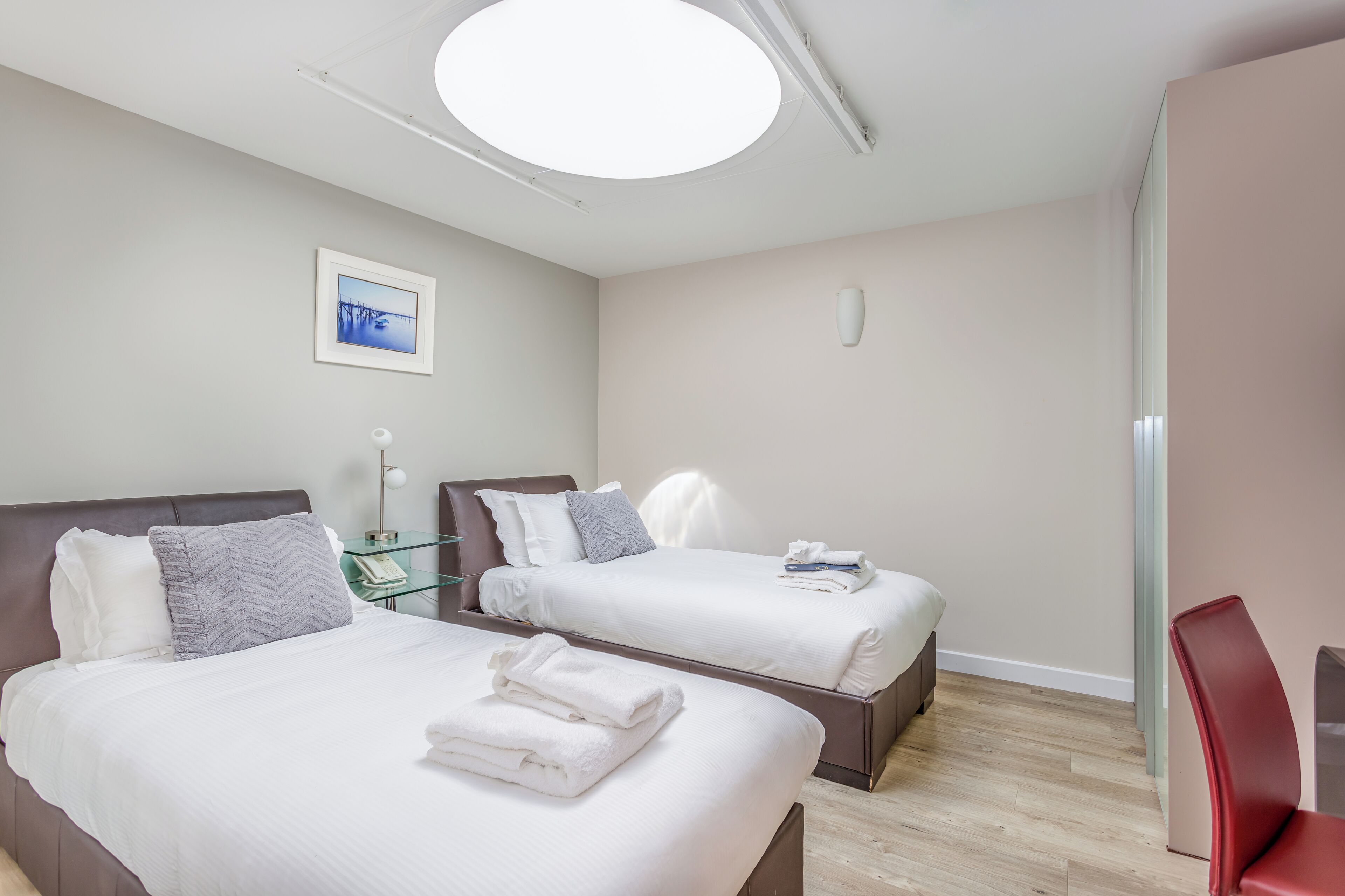 Ein Hotelzimmer mit zwei Betten, einem Nachttisch mit Lampe, einem gerahmten Bild an der Wand und einem roten Sessel.