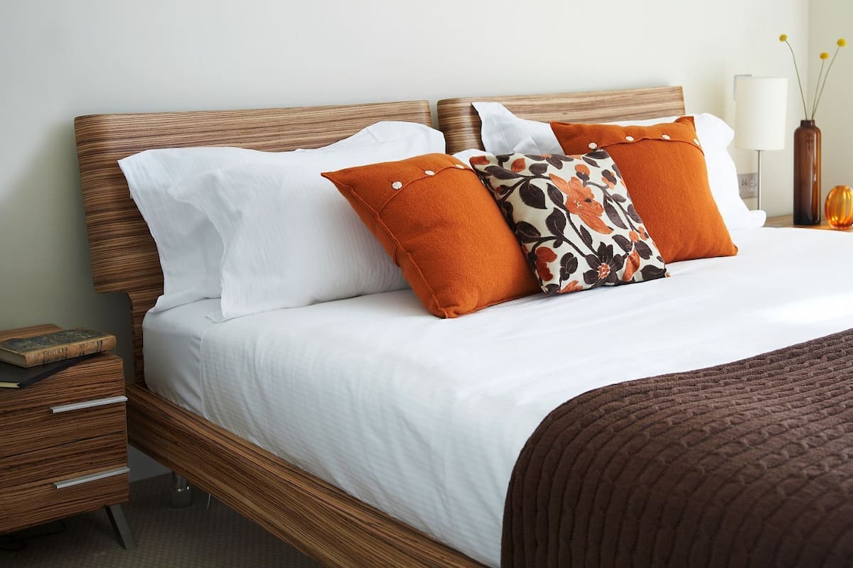 A neatly made bed with white linens, two orange pillows, and a brown throw blanket.