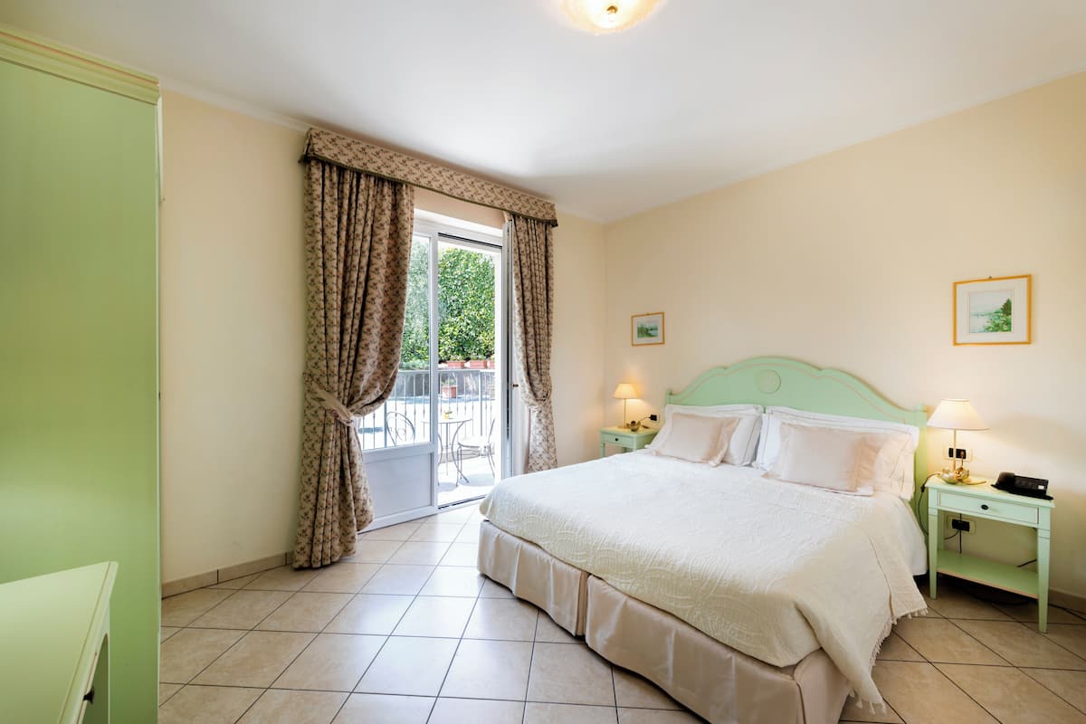 A bedroom with a bed, a nightstand, a lamp, a green wardrobe, and a view of the outdoors through a window with curtains.