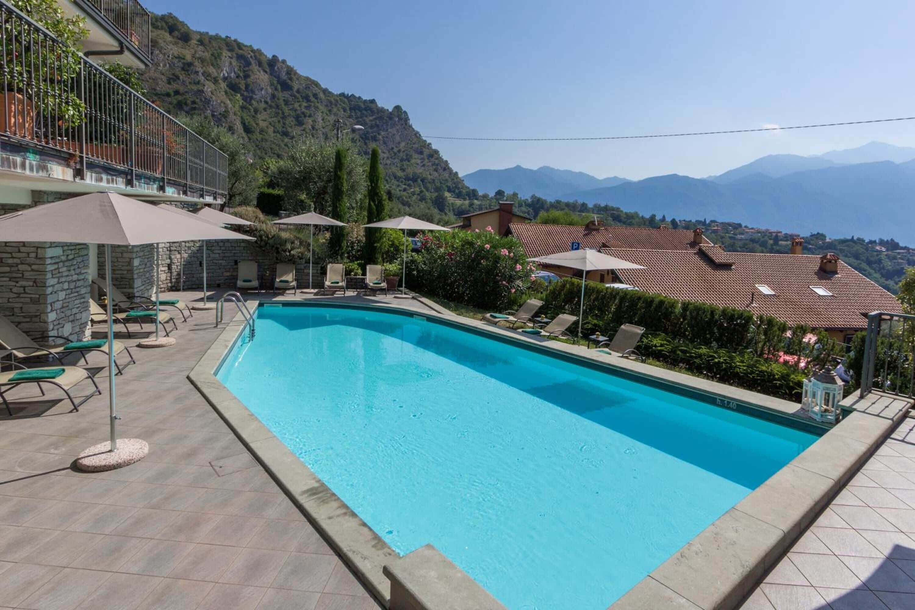 Una piscina con sillas de descanso y sombrillas, rodeada de edificios de piedra y un paisaje montañoso.