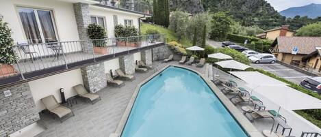 Une piscine entourée de chaises longues et de parasols, le tout bordé par un bâtiment à la façade en pierre et doté d’un balcon.