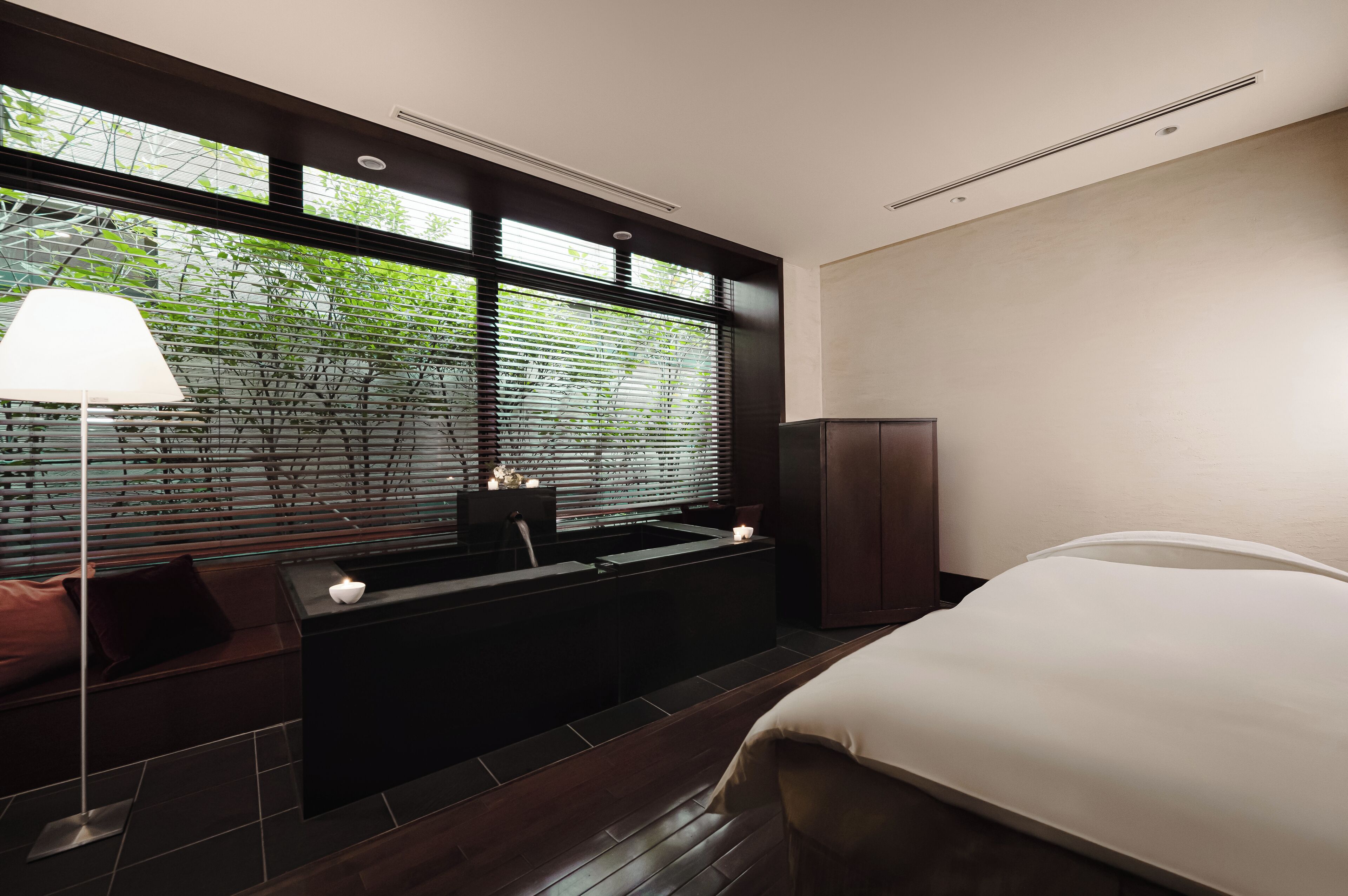 A modern hotel room with a large bed, a built-in bench with a lamp, a small table, and a view through large windows.