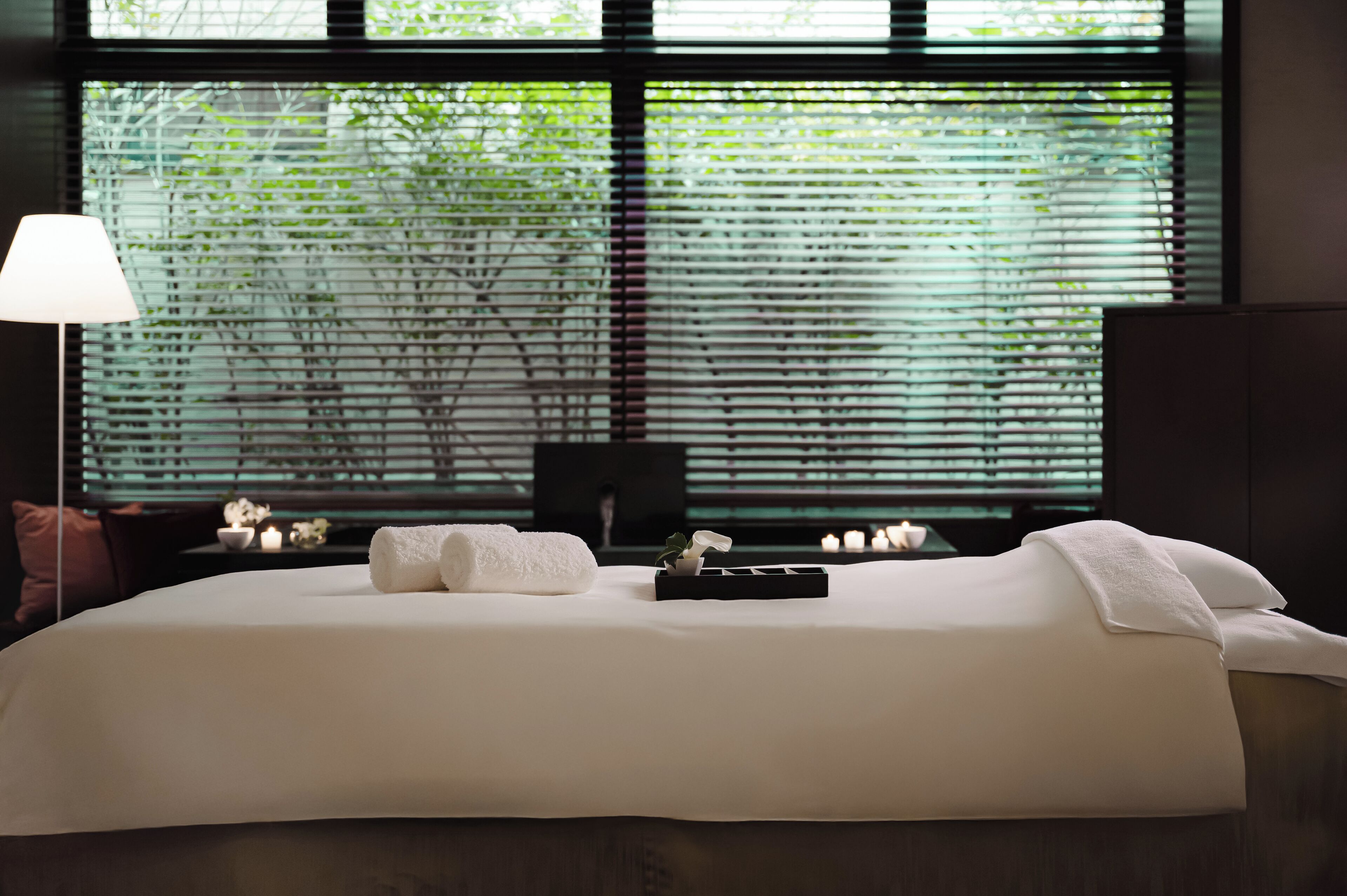 A neatly made bed with pillows and a tray, a bedside lamp, and a window with blinds.
