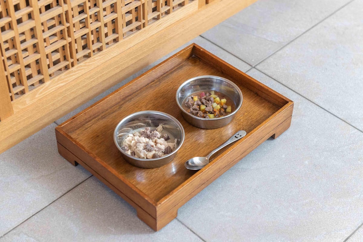 A wooden tray with two metal bowls containing food, placed on a tiled floor.