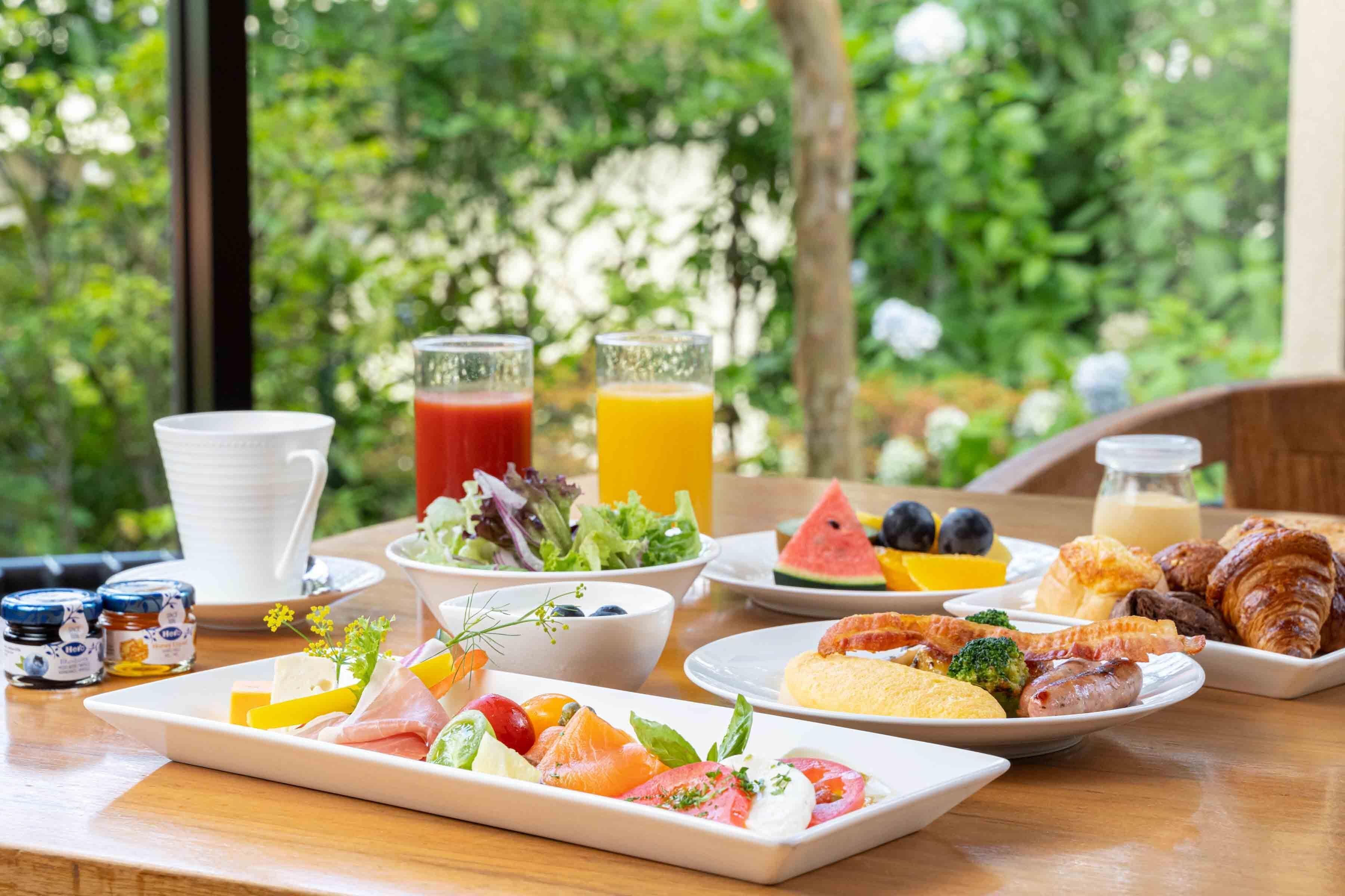 A breakfast table with various food items, including fruits, vegetables, and pastries, set against a backdrop of greenery.