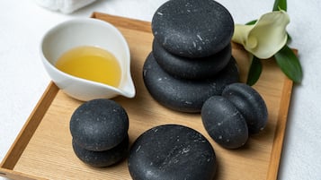A tray with black stones and a small bowl of yellow liquid, possibly oil, on a wooden surface with a white towel and a white flower in the background.