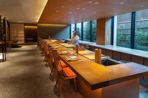 A modern dining area with a long wooden table, multiple chairs, and a chef preparing food.