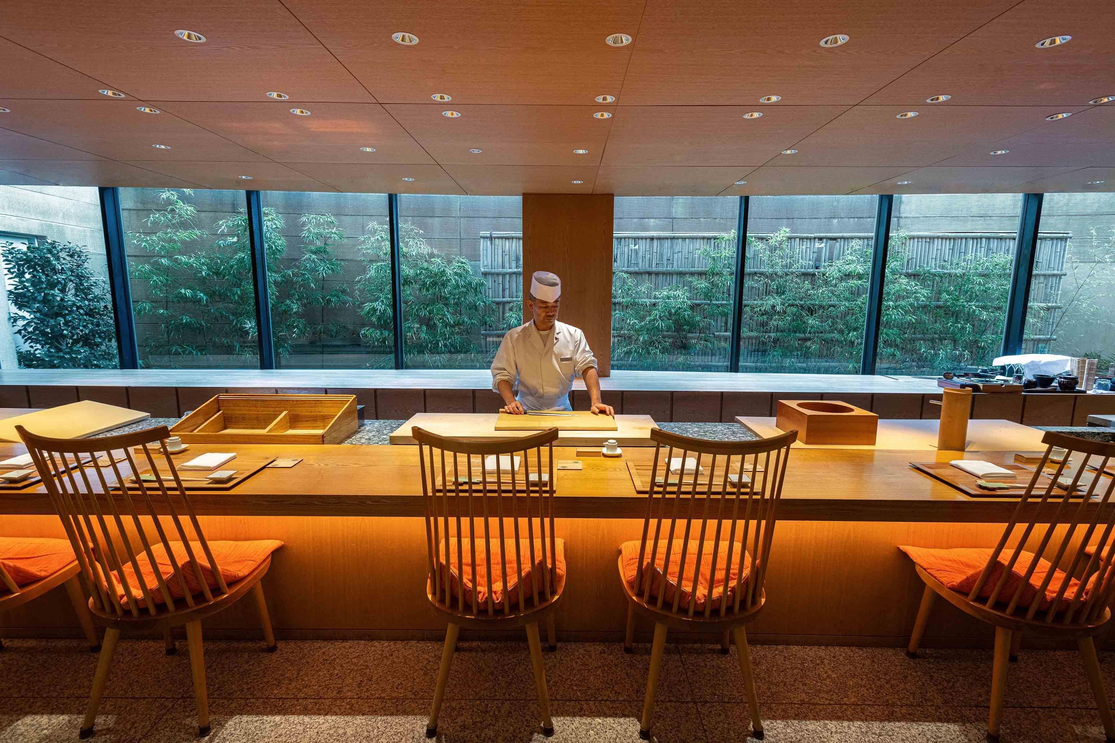 A restaurant with a chef at the counter, large windows, and wooden chairs with orange cushions.