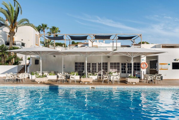 A poolside area with outdoor seating and umbrellas, a clear blue pool, and a white building with windows and a balcony. - Alua Lago Park (Ciutadella de Menorca)