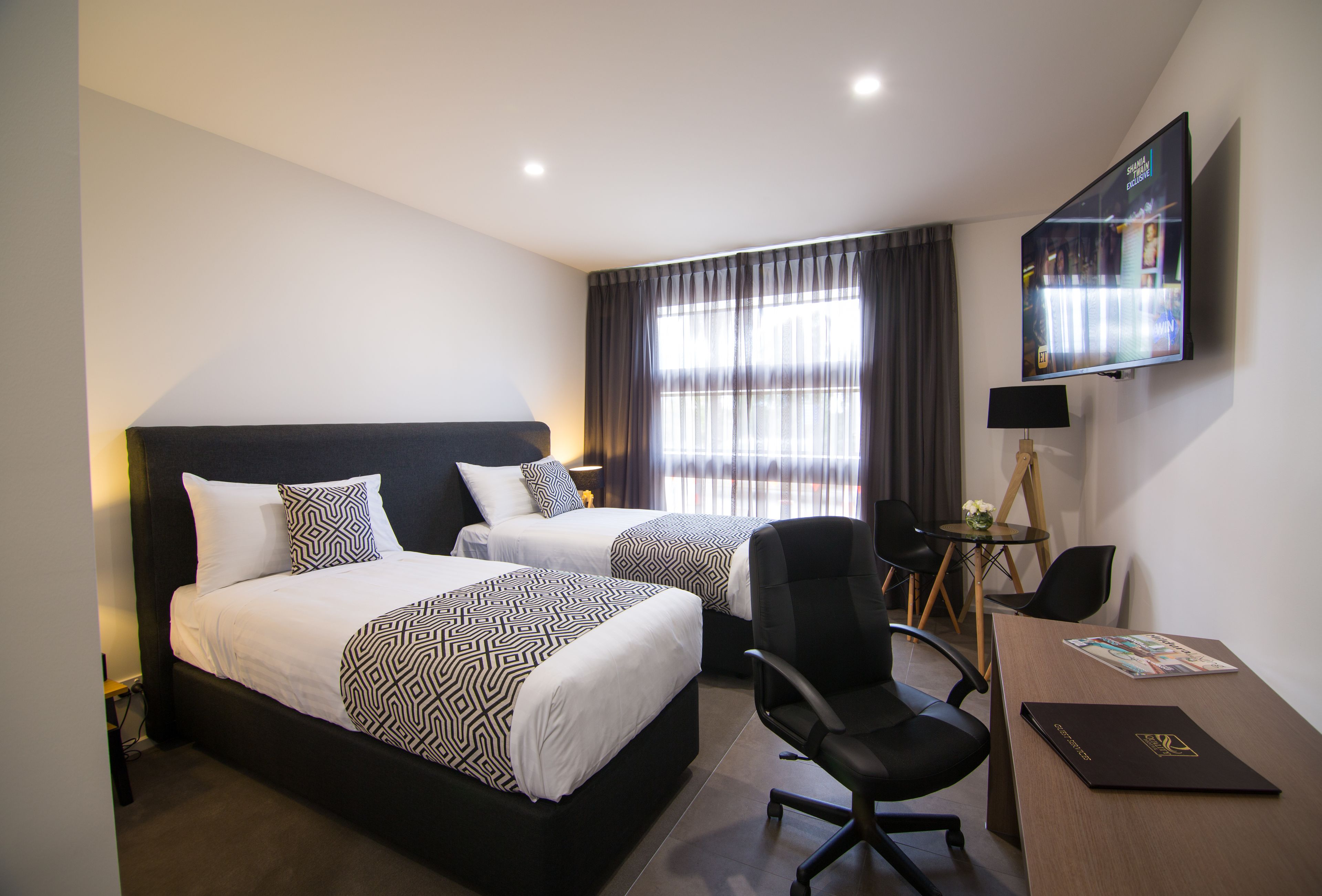 A hotel room with two beds, a desk with a laptop, a chair, a TV, and a window with curtains.