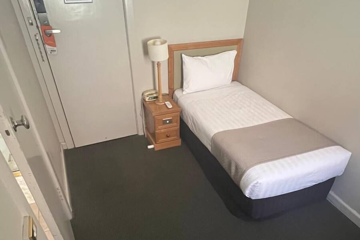 Ein kleines, gemütliches Zimmer mit einem Einzelbett, einem Nachttisch mit Lampe, einem Sessel und einer Tür mit Schloss.