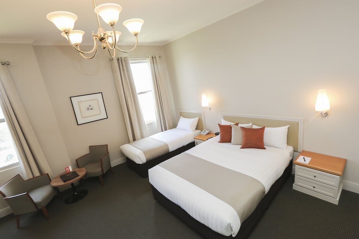 Ein Hotelzimmer mit zwei Einzelbetten, einem Schreibtisch mit Stuhl, einem kleinen Tisch, einem Nachttisch und einer Deckenleuchte.