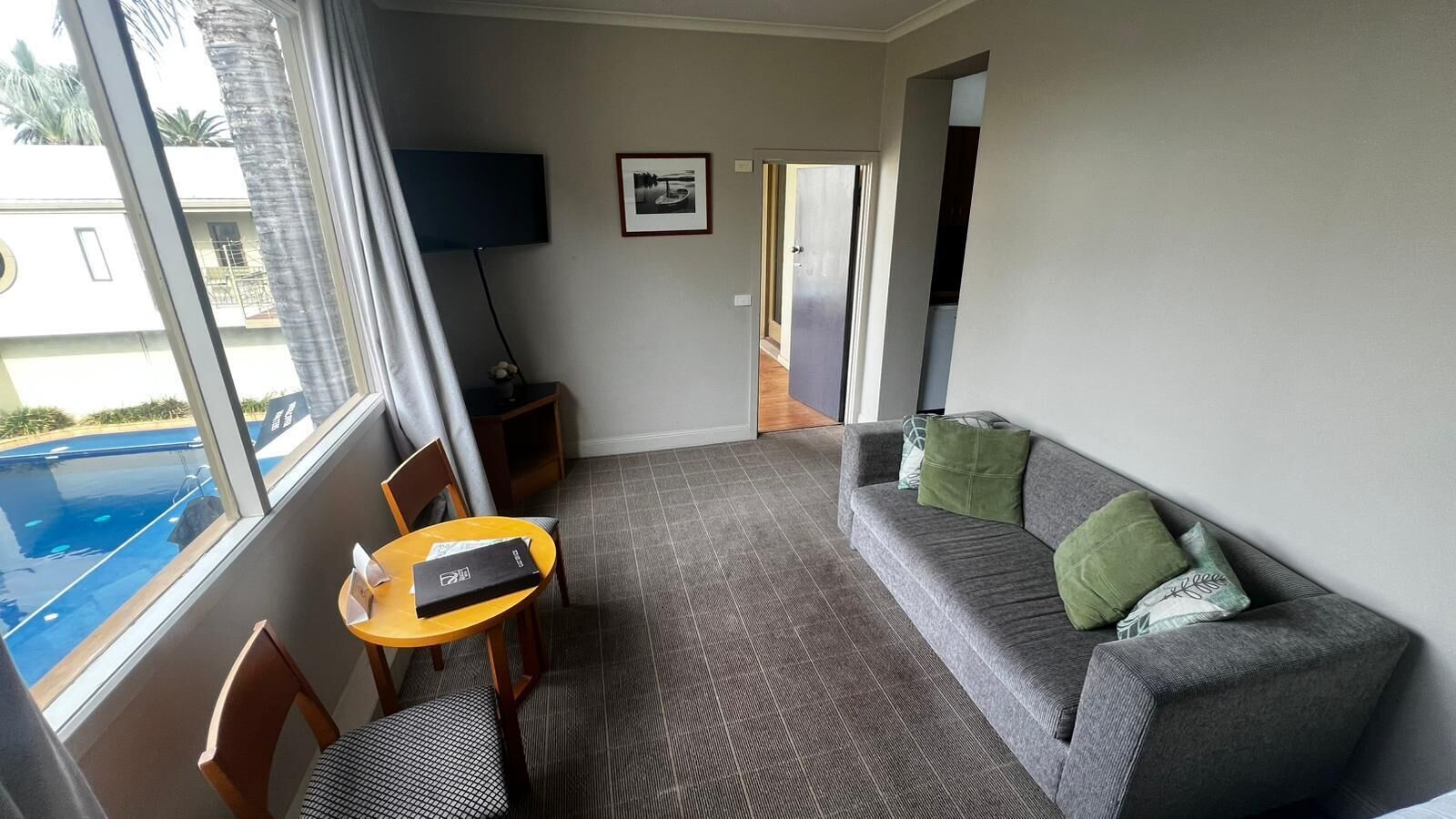 A room with a grey sofa, a small round table, a TV, and a view of a swimming pool.