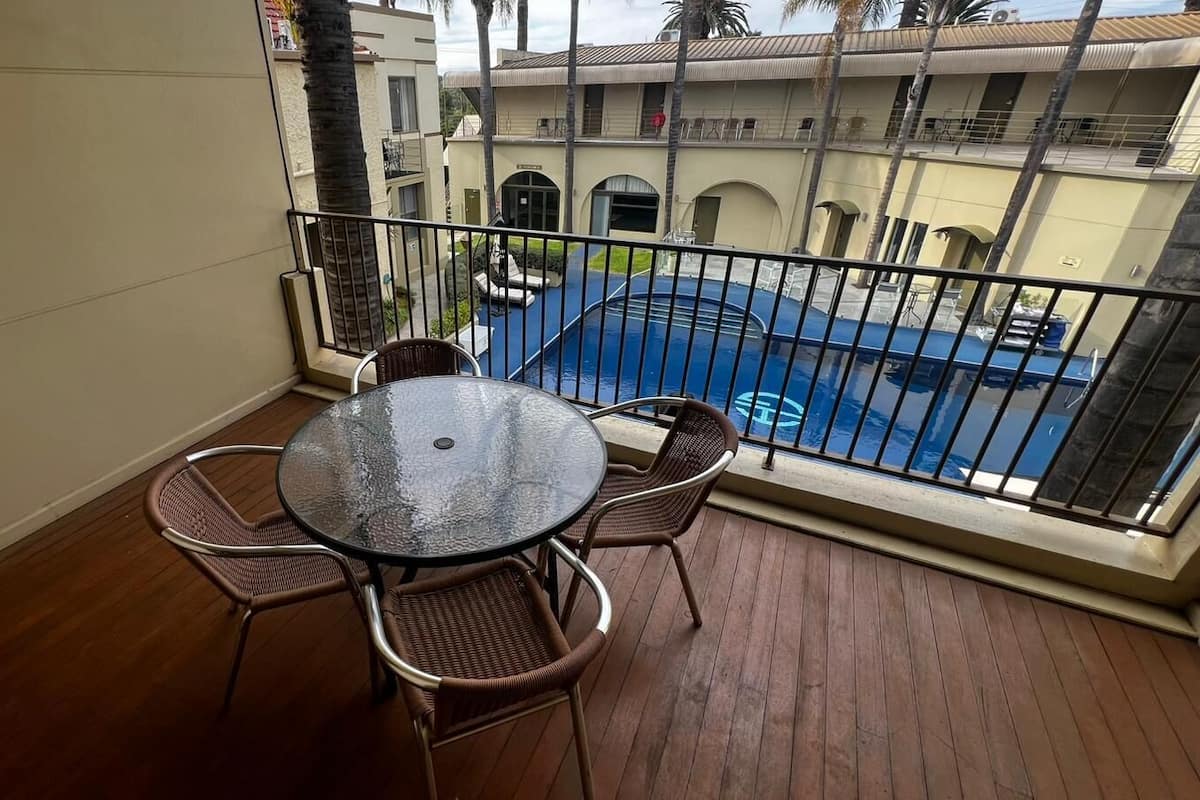 Ein Balkon mit rundem Glastisch und Korbstühlen, der auf einen Swimmingpool und ein Gebäude mit Balkonen blickt.