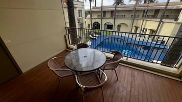 A balcony with a round glass table and wicker chairs, overlooking a swimming pool and a building with balconies.