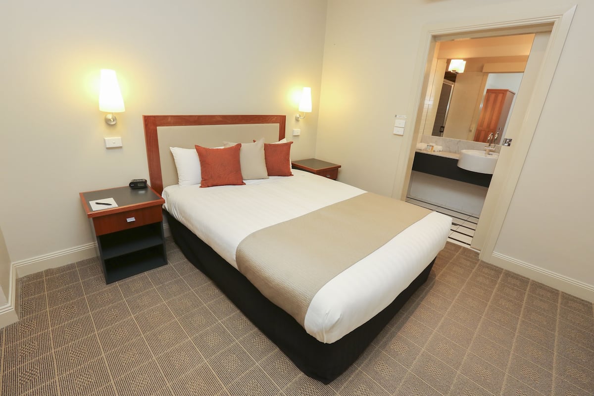 Ein Hotelzimmer mit einem großen Bett, zwei Nachttischen, einem Schreibtischchen, einem Badezimmer mit Waschbecken und Spiegel sowie einem Teppichboden.