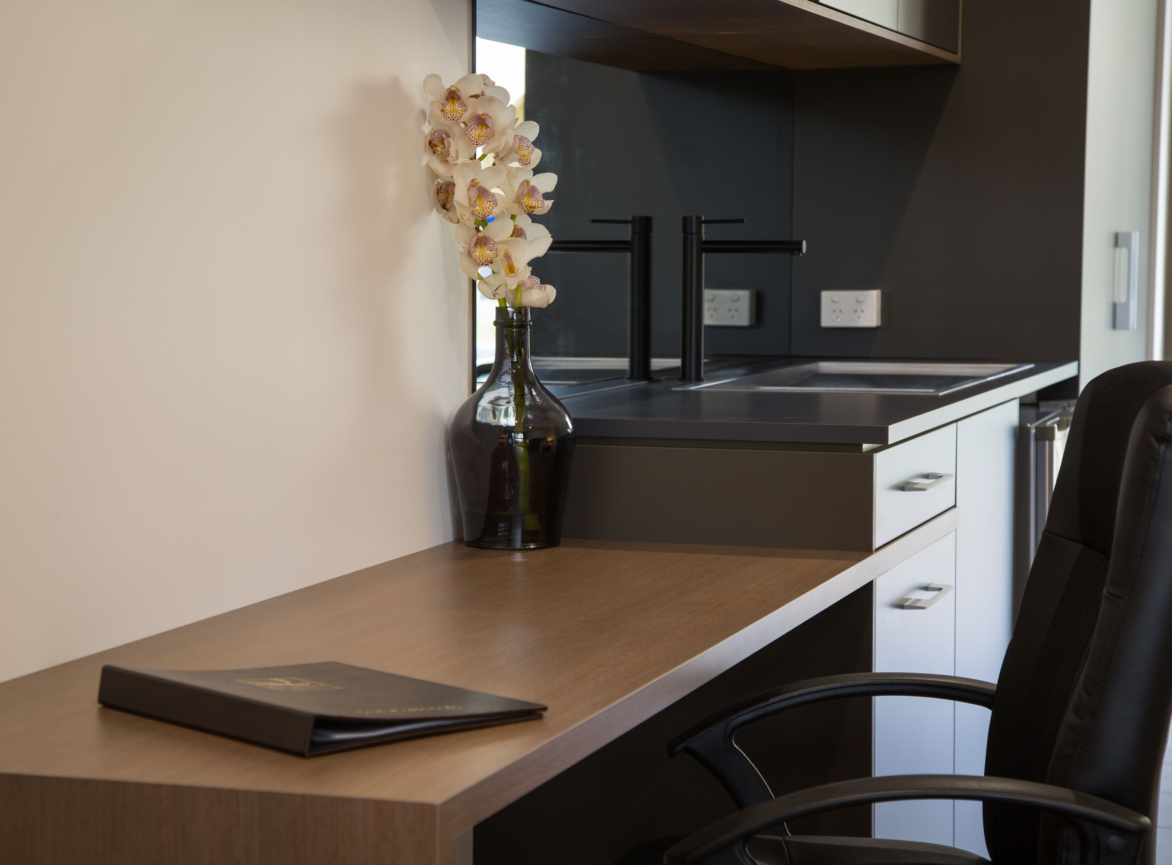 Une cuisine moderne avec un plan de travail foncé, un vase de fleurs blanches, un bureau en bois et une chaise de bureau noire.