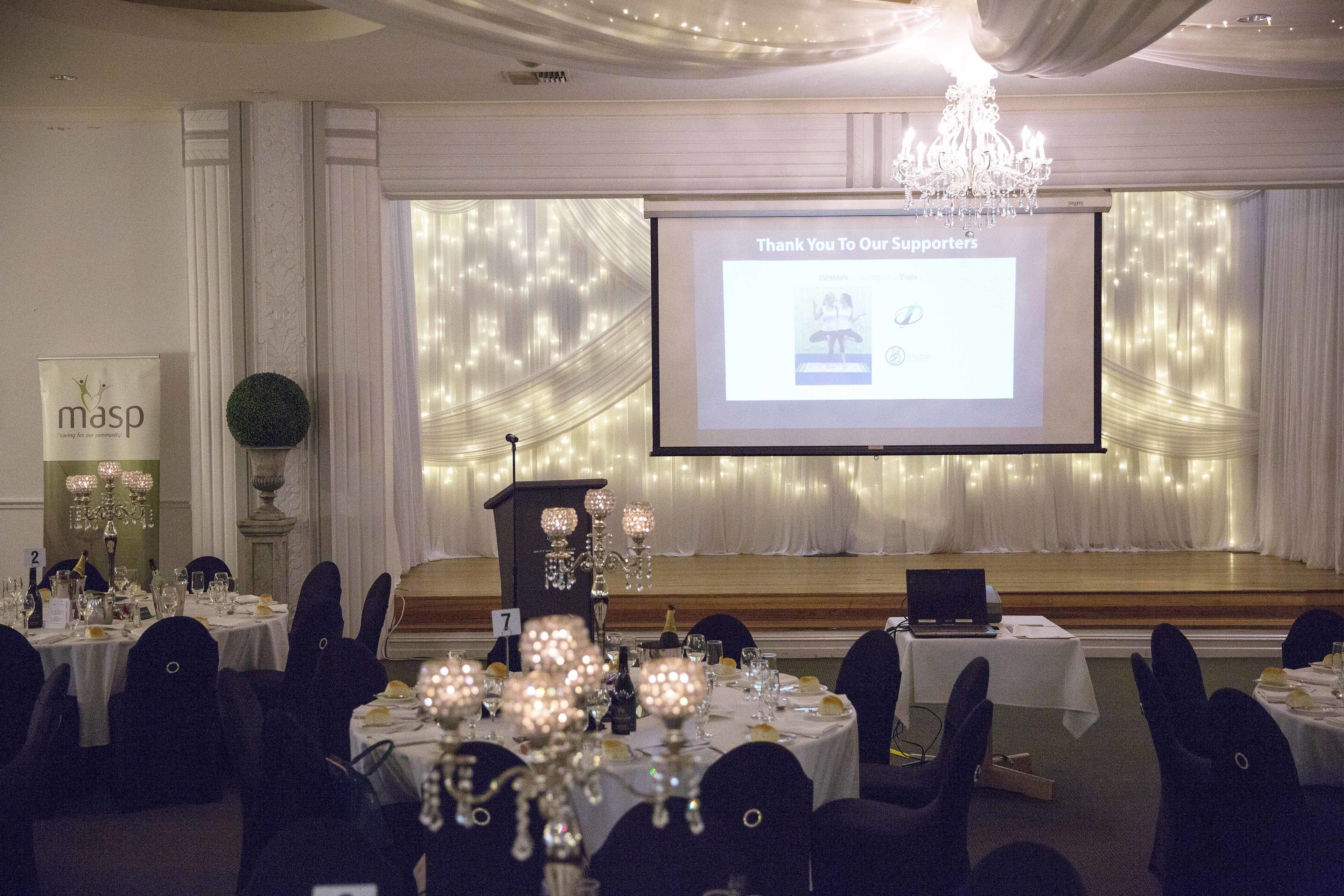 A banquet hall with tables set for a formal event, a projection screen displaying a thank you message, and a chandelier.