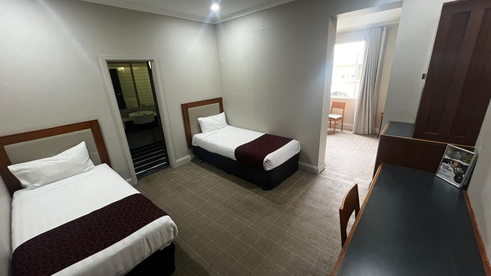 A hotel room with two beds, a desk, a chair, a bathroom with a sink, and a view of the outside.