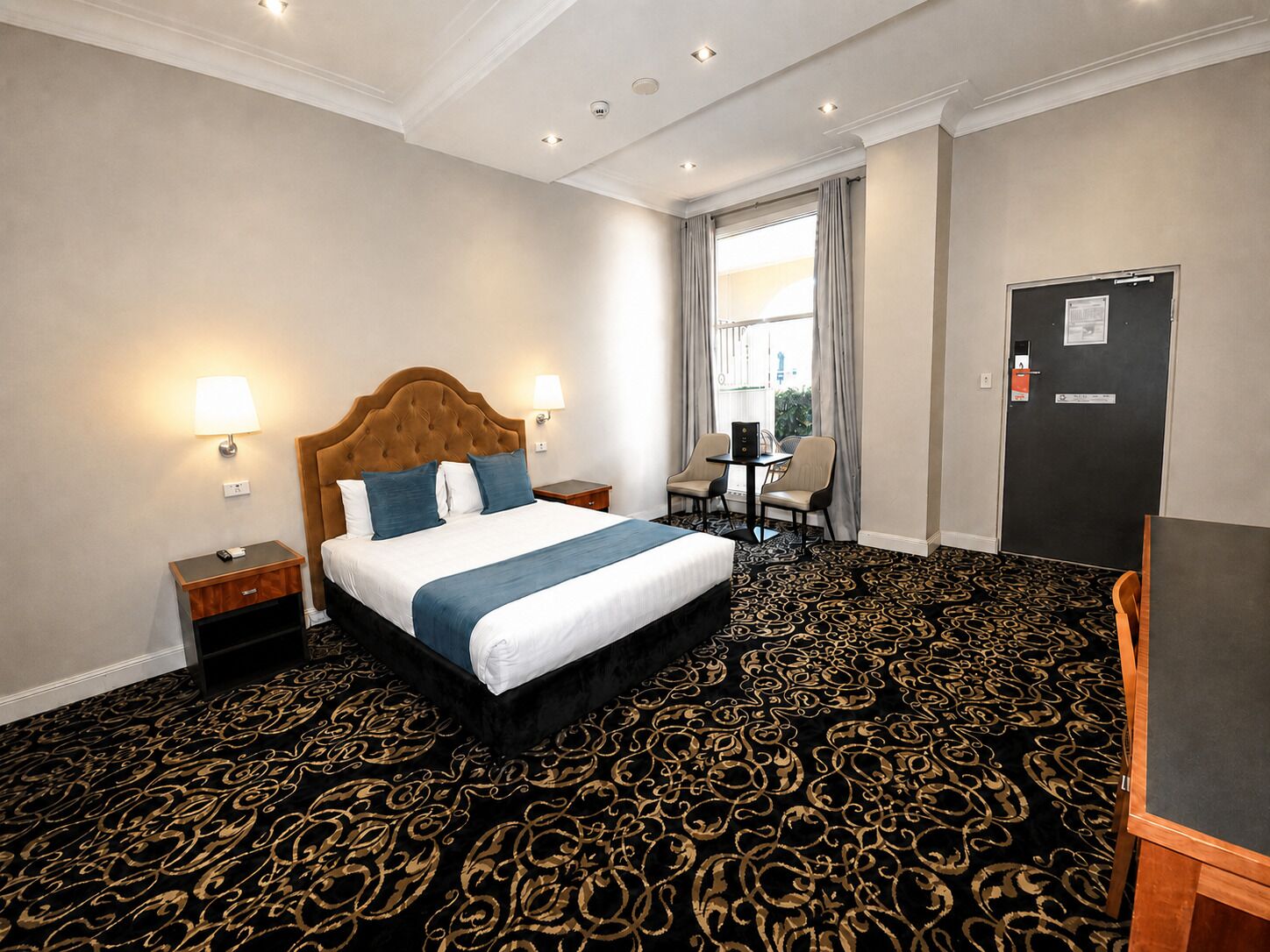 Une chambre d’hôtel avec un grand lit, des tables de chevet, un bureau avec des chaises, une fenêtre avec des rideaux et une porte avec une pancarte.