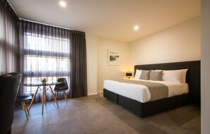 Une chambre d’hôtel moderne équipée d’un lit, d’un bureau, de deux chaises, d’une fenêtre avec des rideaux et d’un tableau accroché au mur.