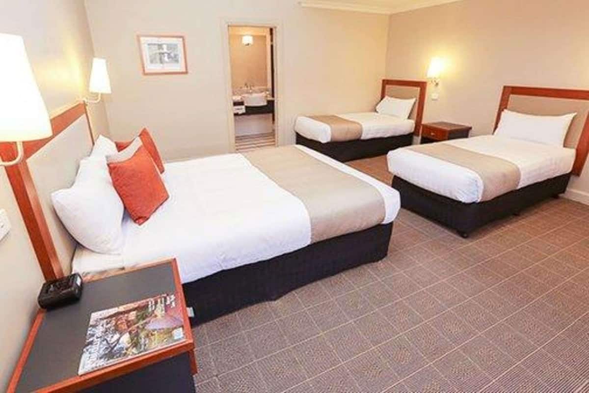Une chambre d’hôtel avec trois lits, une petite table sur laquelle se trouve un magazine, et une salle de bain visible à travers une porte ouverte.