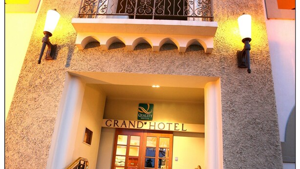 Het uiterlijk van het Grand Hotel, met een opvallende ingang, een balkon met decoratieve hekwerk en twee aan de muur bevestigde lampen.