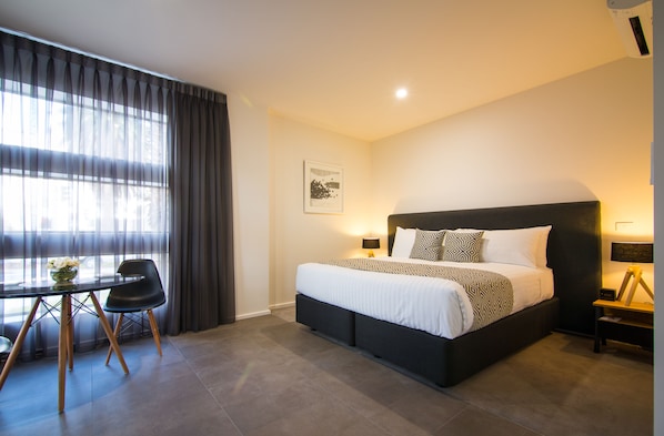 A modern hotel room with a large bed, a desk with a chair, and a view through sheer curtains.
