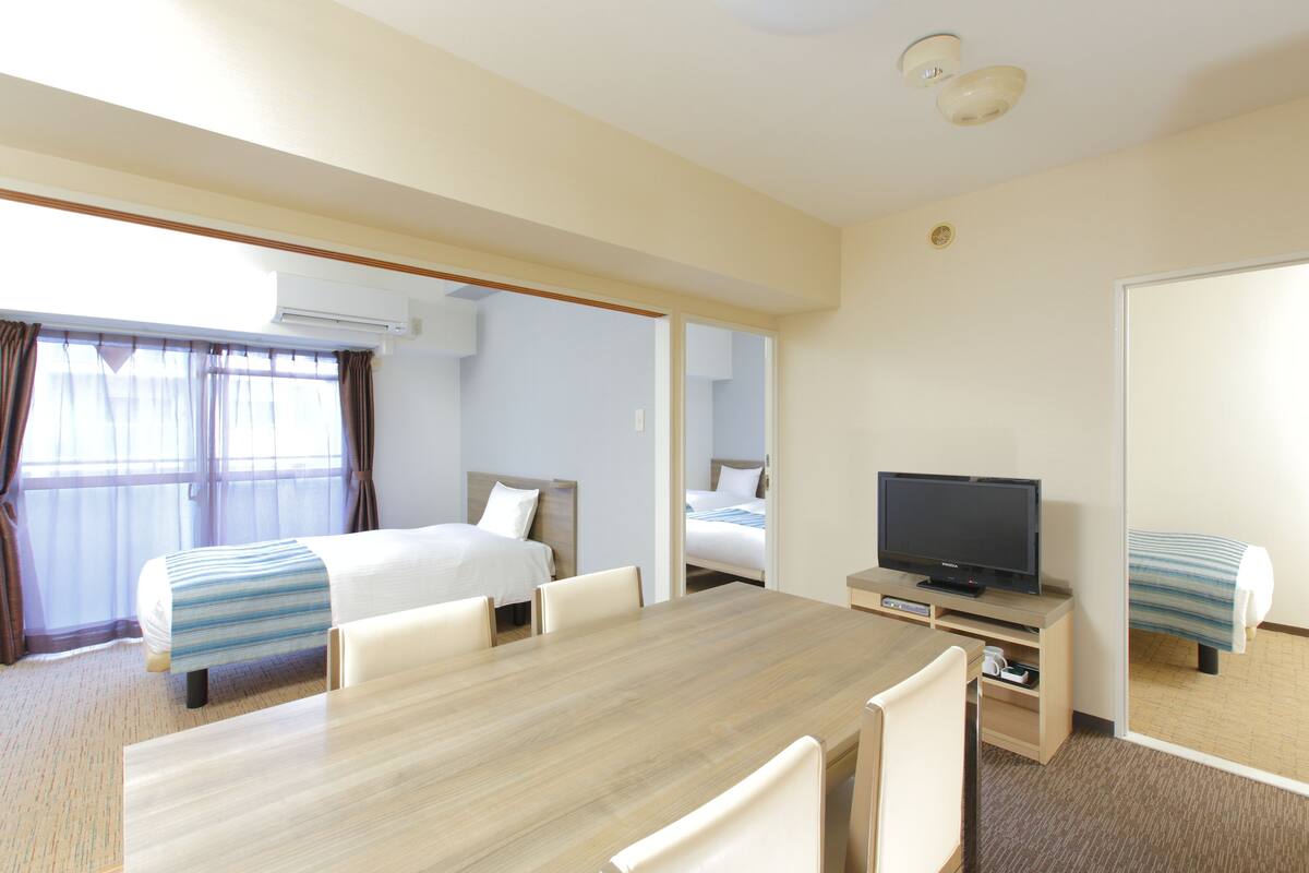 Habitación de hotel con una mesa de comedor de madera, dos camas individuales, un televisor y un equipo de aire acondicionado.