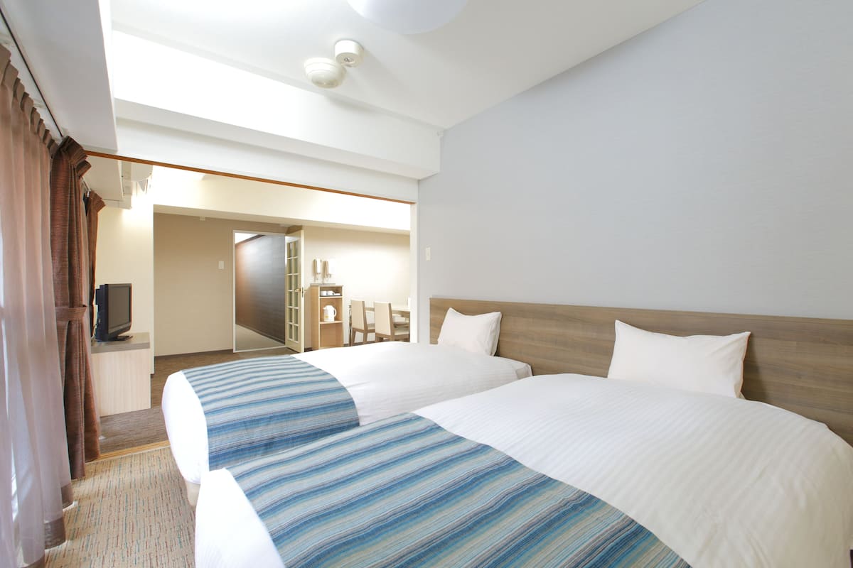 Habitación de hotel con dos camas, televisión, escritorio y baño visible a través de una puerta abierta.