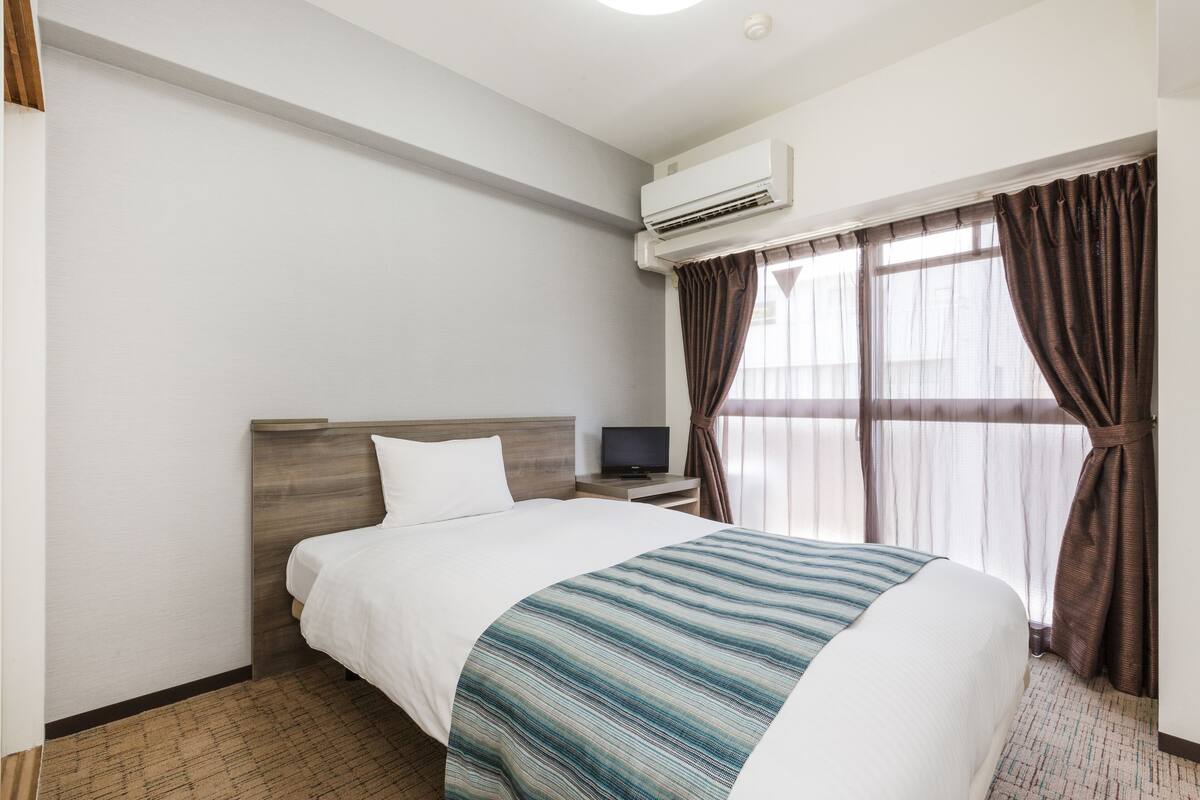Habitación de hotel con cama, televisor en una mesita de noche, aire acondicionado y ventana con cortinas.