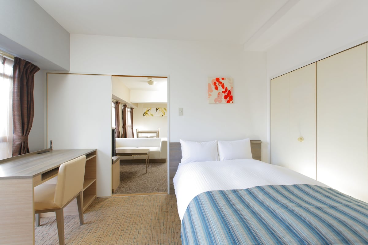 Habitación de hotel con cama, escritorio y silla. Hay un espejo en la pared y una puerta que da a otra habitación.