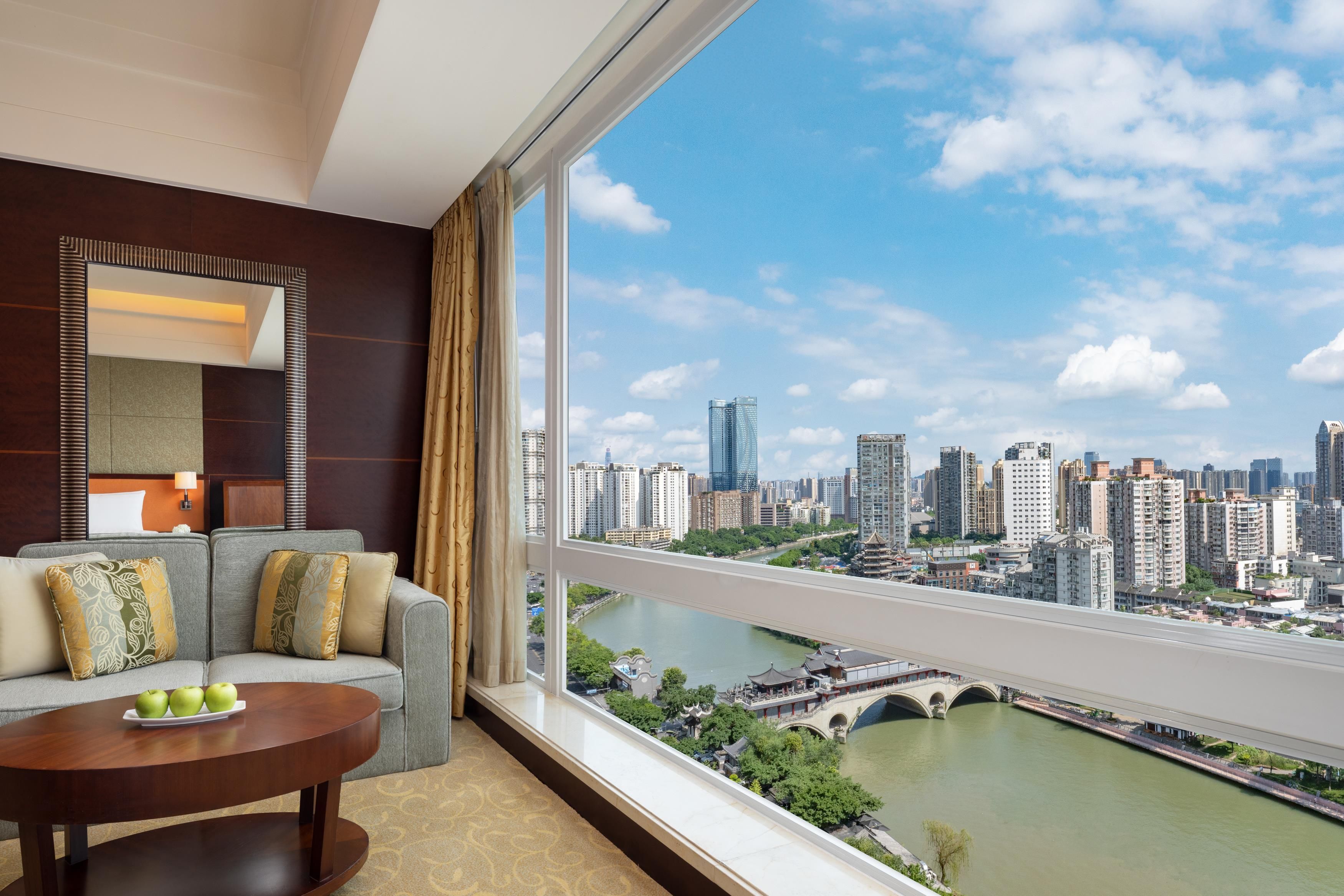 A hotel room with a large window, a sofa, a coffee table, and a view of a cityscape and river.
