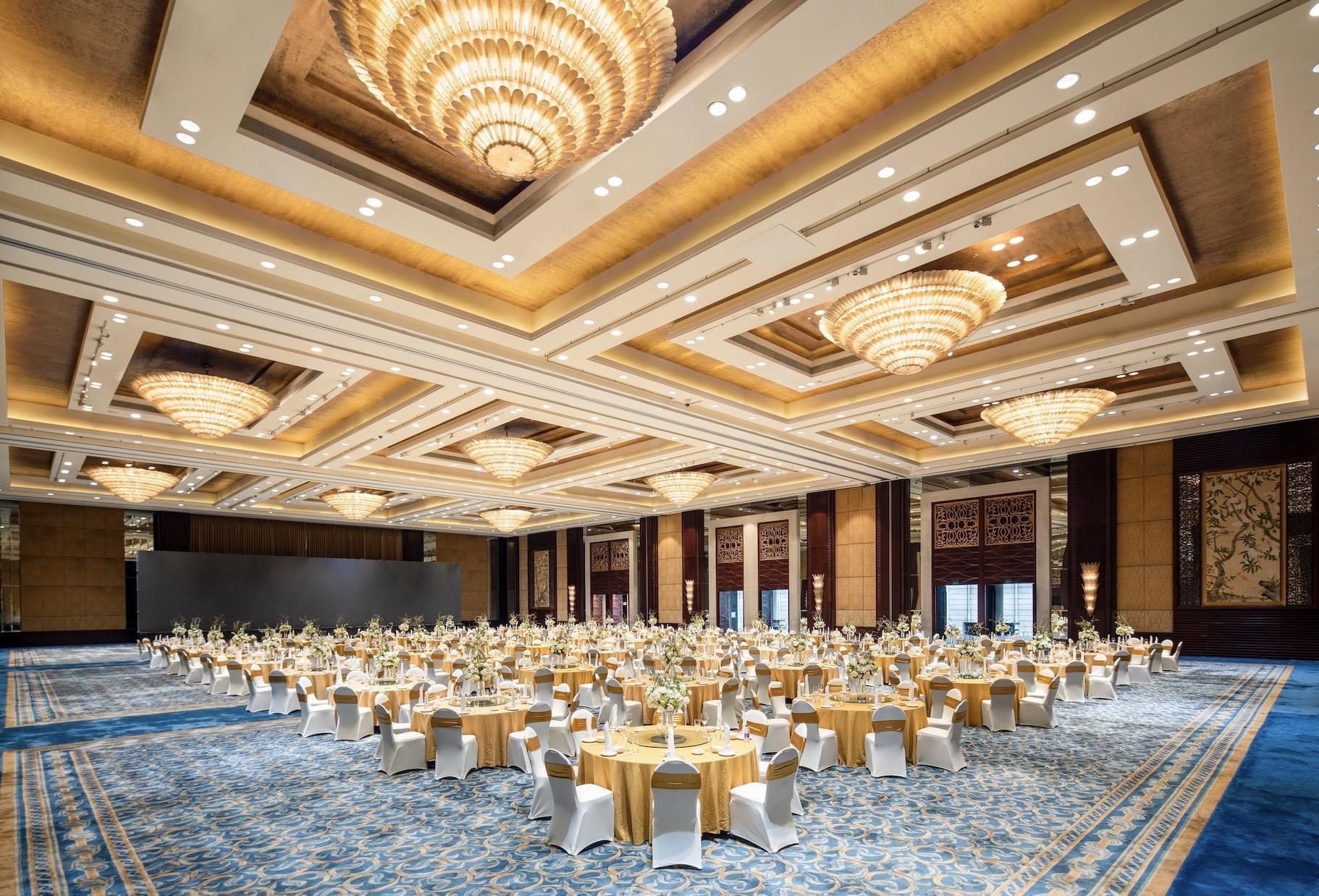 Un amplio salón de banquetes con mesas redondas, sillas blancas y cintas doradas, con una alfombra estampada en azul y lujosos candelabros.