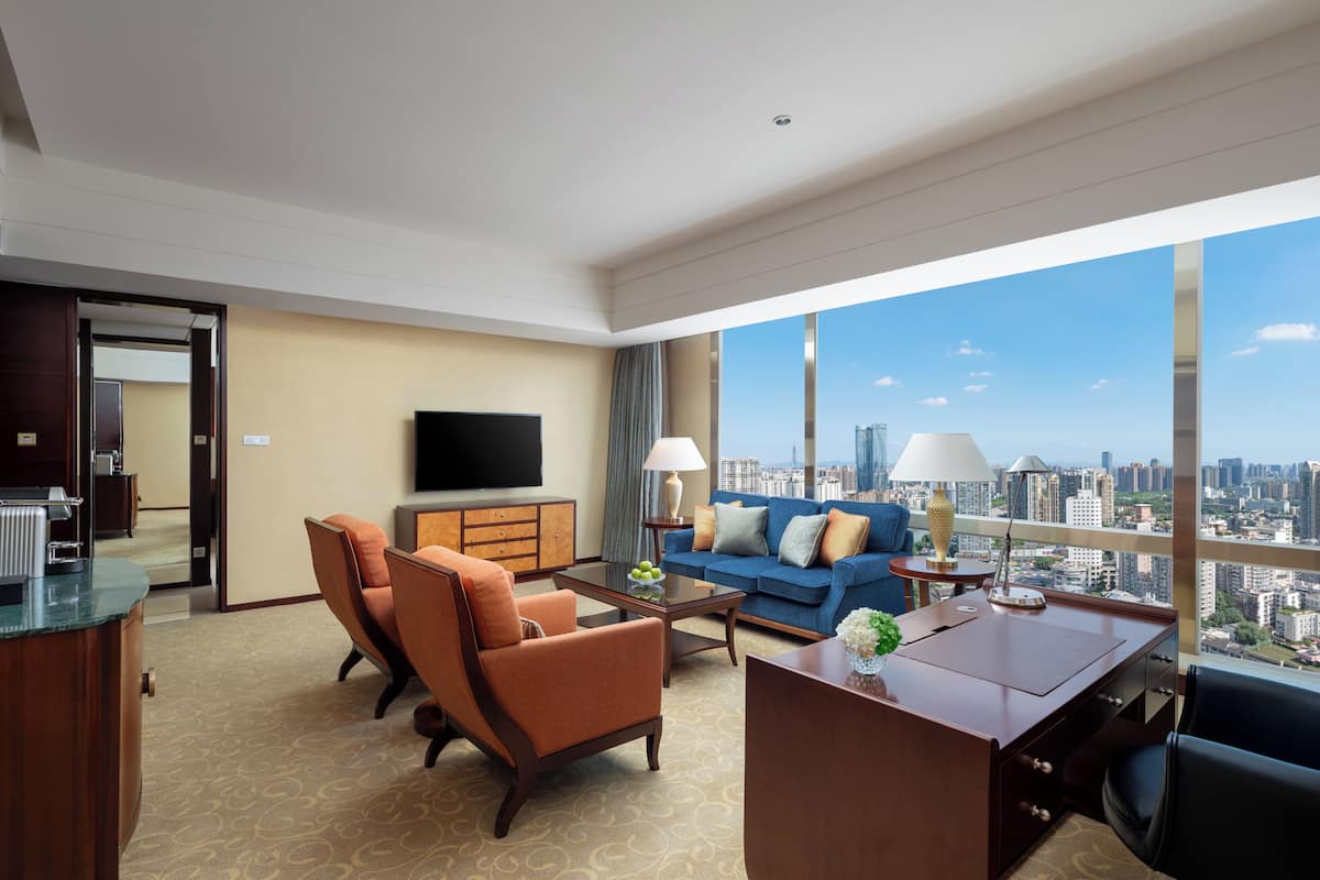 Una habitación de hotel moderna con una amplia vista de la ciudad, un televisor de pantalla plana, un sofá, dos sillones, una mesa de centro y un escritorio.