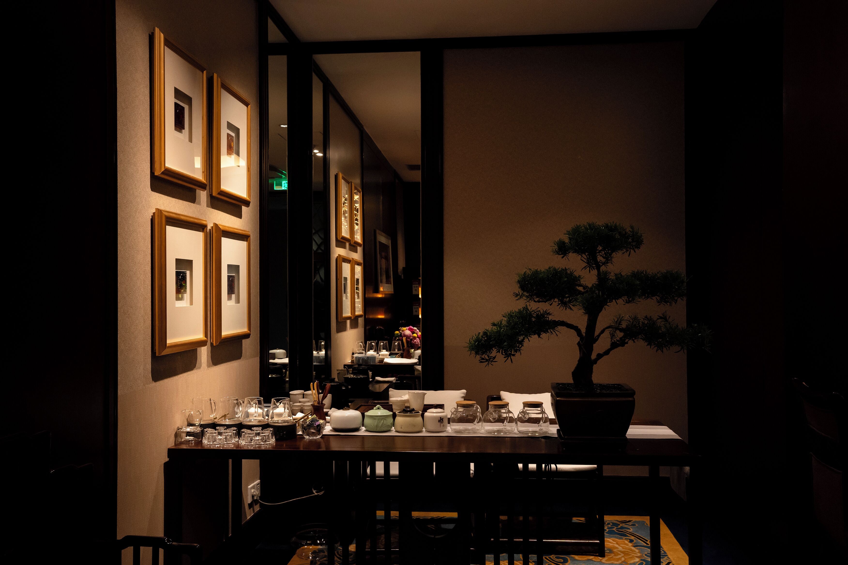 Un comedor tenuemente iluminado con una mesa dispuesta para dos, fotografías enmarcadas en la pared y un bonsái.