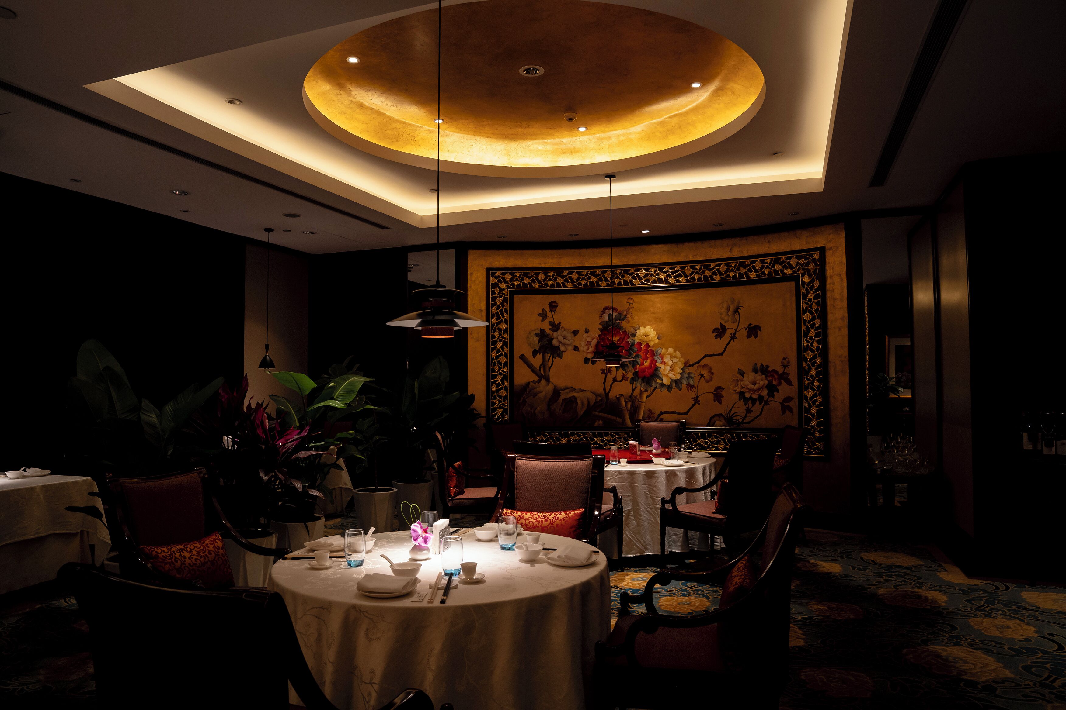 A dimly lit dining area with round tables, chairs, and a large floral painting on the wall.