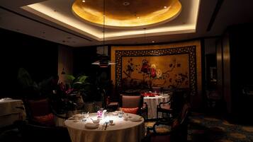 A dimly lit dining area with round tables, chairs, and a large floral painting on the wall.