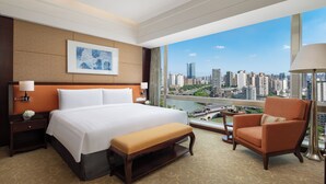A hotel room with a large bed, a chair, a bench, a nightstand, a lamp, and a view of a cityscape. - Shangri-La Chengdu (Chengdu)